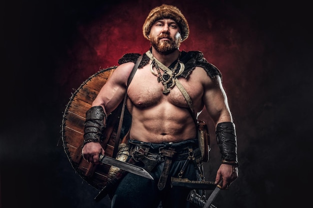 Un vikingo serio vestido con una armadura ligera con un escudo detrás de la espalda sostiene una espada y un hacha. Posando sobre un fondo oscuro con luz roja