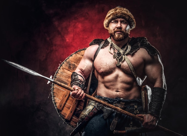 Un vikingo serio vestido con una armadura ligera con un escudo detrás de la espalda sostiene una lanza. Posando sobre un fondo oscuro con luz roja