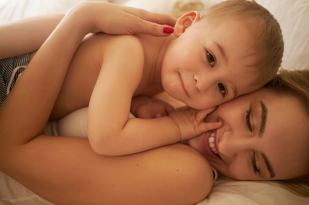 Vínculos familiares y concepto de maternidad. Imagen recortada de la hermosa joven mamá europea relajándose en el dormitorio acostado en sábanas blancas con su adorable hijo, abrazándolo fuerte y sonriendo felizmente