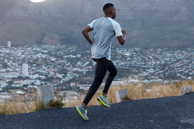 La vista de acción del corredor masculino cubre larga distancia, vestido con leggings casuales y camiseta, posa sobre la vista de las montañas en la carretera, tiene zapatos deportivos, recupera el aliento durante el entrenamiento cardiovascular. Movimiento, concepto de velocidad