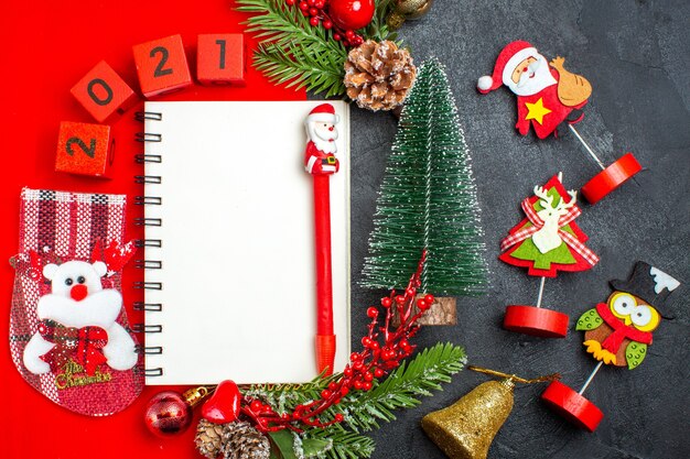 Vista aérea de accesorios de decoración de cuaderno espiral números de calcetín navideño de ramas de abeto en una servilleta roja y árbol de Navidad sobre fondo oscuro
