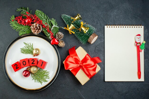 Vista aérea de los accesorios de decoración de números en una placa, ramas de abeto, cono de coníferas, árbol de navidad, cuaderno con lápiz sobre fondo oscuro