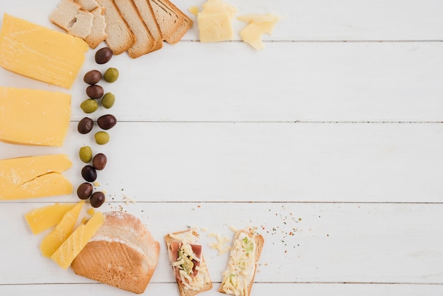 Una vista aérea de las aceitunas; Rebanada de queso y pan en mesa de madera blanca