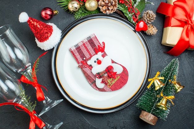 Vista aérea del calcetín de Navidad en el plato de cena árbol de navidad ramas de abeto cono de coníferas caja de regalo sombrero de santa claus copas de vidrio caído sobre fondo negro