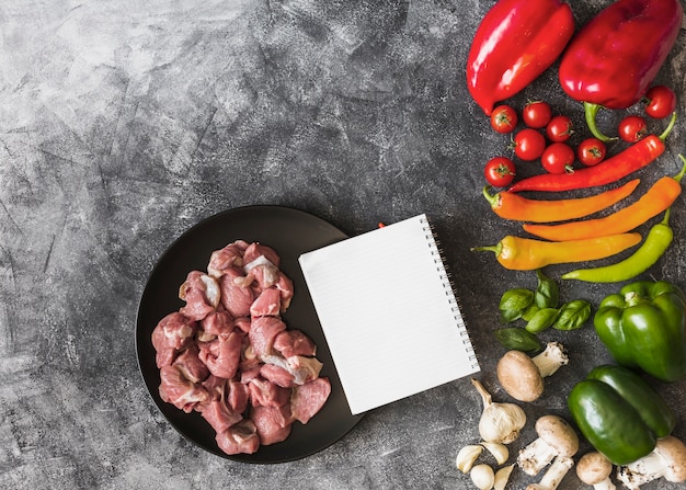 Una vista aérea de carne cruda con cuaderno y verduras de colores sobre fondo manchado