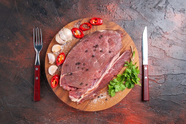 Vista aérea de la carne roja en bandeja de madera y ajo, limón, pimienta, cebolla, tenedor y cuchillo sobre fondo oscuro