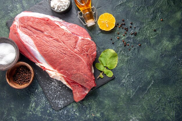 Vista aérea de la carne roja cruda fresca en bandeja negra pimienta limón botella de aceite caído sobre fondo de color oscuro