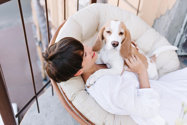 Vista aérea de una chica morena bronceada mirando con una sonrisa al perro beagle soñoliento sentado al lado