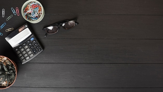 Foto gratuita una vista aérea de clips de papel de colores; calculadora y gafas en mesa de madera negra