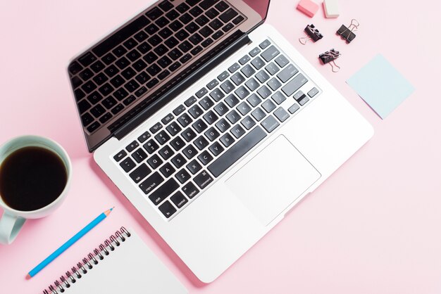Una vista aérea de una computadora portátil abierta; taza de café; lápiz; Bloc de notas y clip sobre fondo rosa