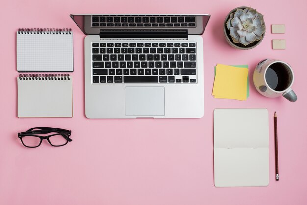 Una vista aérea de la computadora portátil; bloc; los anteojos; notas adhesivas; Planta de cactus y taza de té sobre fondo rosa