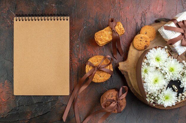 Vista aérea de un cuaderno cerrado y una caja de regalo bellamente empaquetada flor y anillo en una caja en forma de corazón en una tabla de madera galletas apiladas en un fondo de color mixto