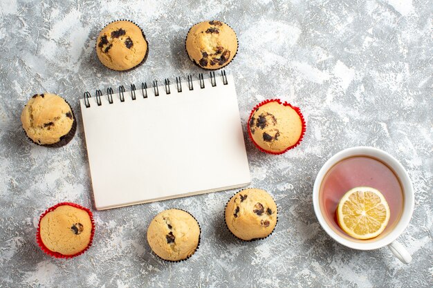 Vista aérea del cuaderno cerrado entre deliciosos cupcakes pequeños con chocolate y mano sosteniendo una taza de té negro con limón sobre la superficie del hielo