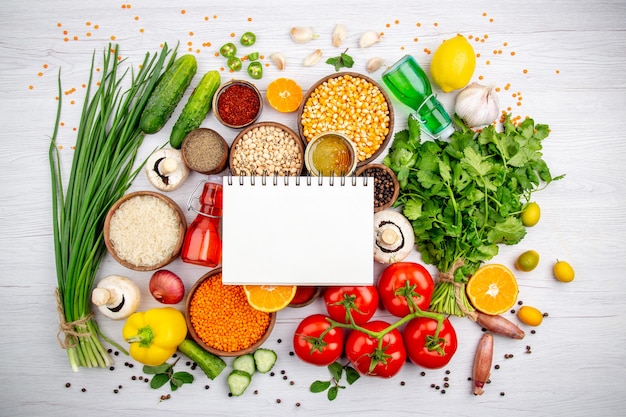 Vista aérea de un cuaderno de espiral sobre verduras frescas limón granos de maíz limón botella de aceite caído miel sobre fondo blanco.