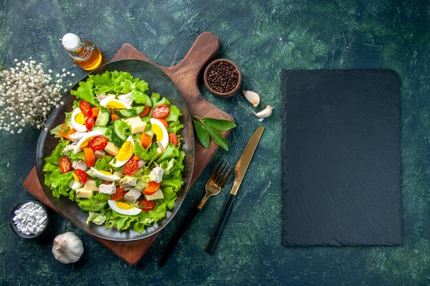 Vista aérea de la deliciosa ensalada con ingredientes frescos en la tabla de cortar de madera, especias, botella de aceite, cubiertos de ajos, sobre fondo negro de colores de mezcla