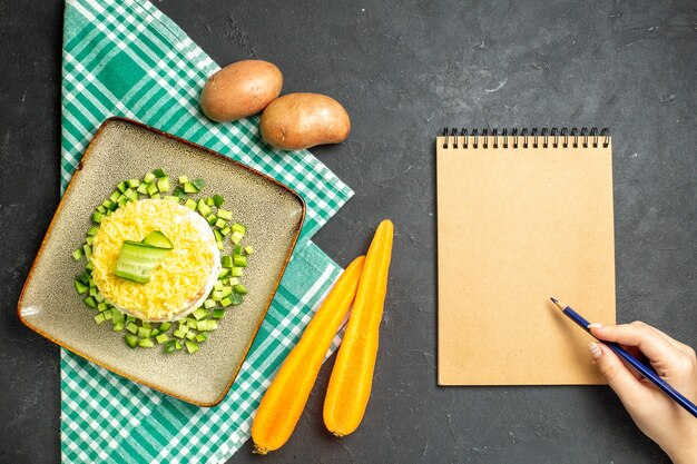 Vista aérea de la deliciosa ensalada servida con pepino picado en zanahorias y papas de toalla verde a la mitad dobladas junto al cuaderno sobre fondo oscuro