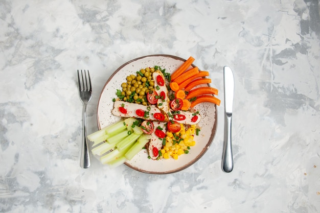 Vista aérea de una deliciosa ensalada con varios ingredientes en un plato y cubiertos en una superficie blanca con espacio libre