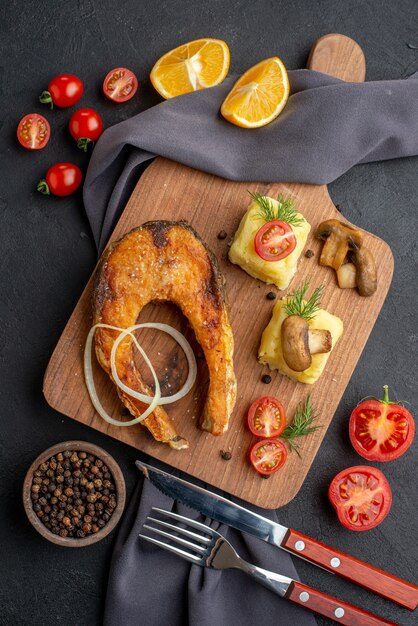 Vista aérea de la deliciosa harina de pescado frito con champiñones tomates queso sobre tabla de madera rodajas de limón pimienta sobre cubiertos de toalla de color oscuro en superficie negra