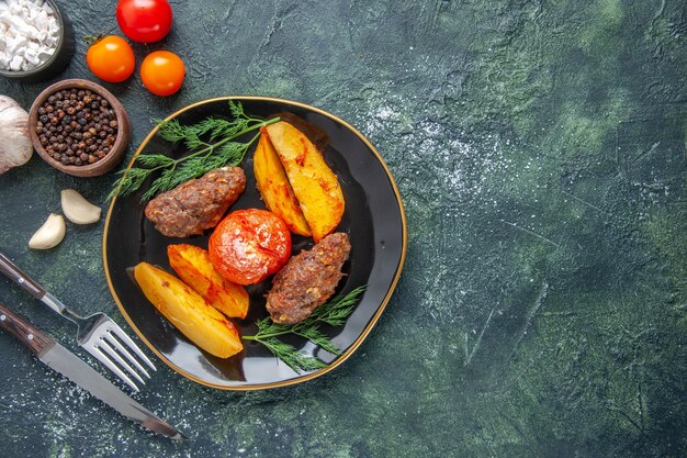 Vista aérea de deliciosas chuletas de carne al horno con patatas y tomates en un plato negro cubiertos, especias, ajo, tomates en el lado derecho