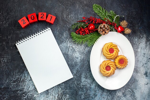 Vista aérea de deliciosas galletas en un plato blanco y adornos de año nuevo números de sombrero de santa claus siguiente cuaderno sobre superficie oscura