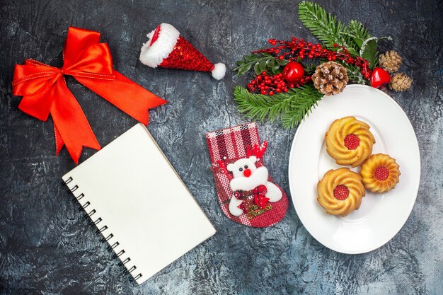 Vista aérea de deliciosas galletas en un plato blanco y adornos sombrero de santa claus cinta roja calcetín de año nuevo junto al cuaderno sobre una superficie oscura