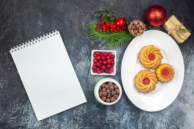 Vista aérea de deliciosas galletas en un plato blanco y decoraciones de año nuevo cornel de regalo en una olla pequeña y un cuaderno sobre una superficie oscura