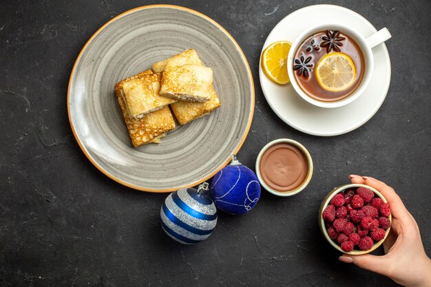 Vista aérea de deliciosos panqueques frescos en un plato blanco y una taza de té negro, chocolate, frambuesa, accesorios de decoración sobre fondo oscuro