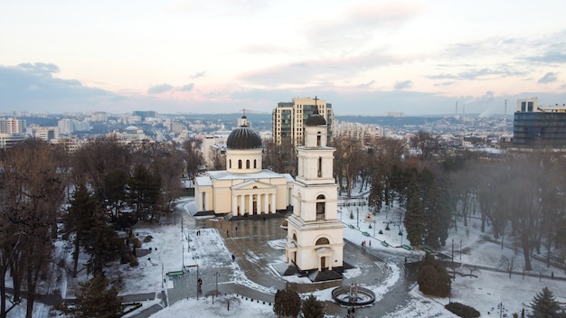 Vista aérea de drone del centro de Chisinau en invierno. Vista panorámica del parque central con nieve, árboles y varias personas que caminan, campanario, catedral, edificios en el fondo.