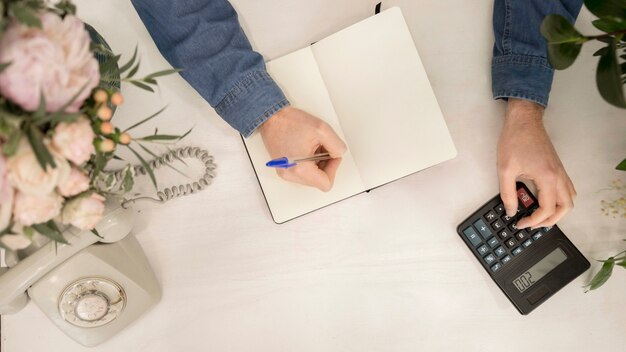 Una vista aérea de la escritura de la florista en el cuaderno usando la calculadora en el escritorio blanco