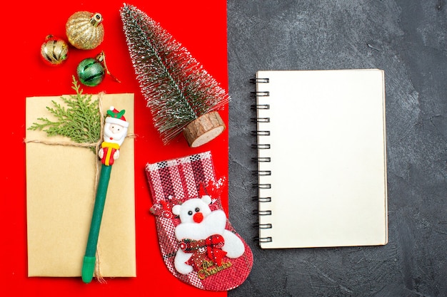 Vista aérea del estado de ánimo navideño con accesorios de decoración del árbol de navidad calcetín de regalo junto al cuaderno sobre fondo rojo y negro