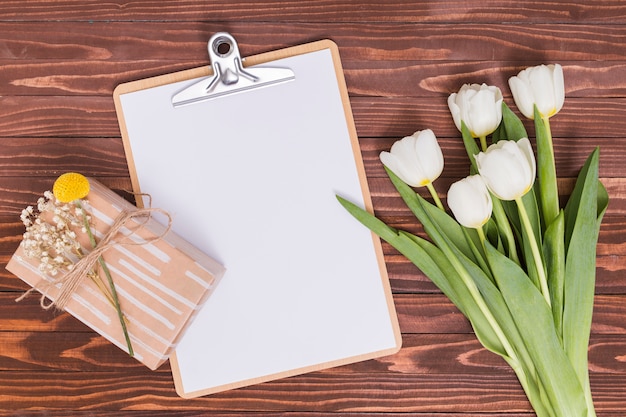 Vista aérea de flores de tulipán blanco; Hoja en blanco; Portapapeles y caja de regalo sobre escritorio de madera.