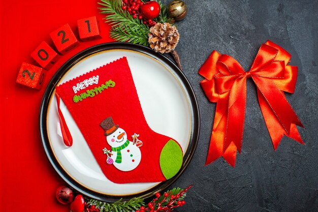 Vista aérea del fondo de año nuevo con calcetín de Navidad en accesorios de decoración de plato de cena ramas de abeto y números en una servilleta roja y cinta roja sobre una mesa negra