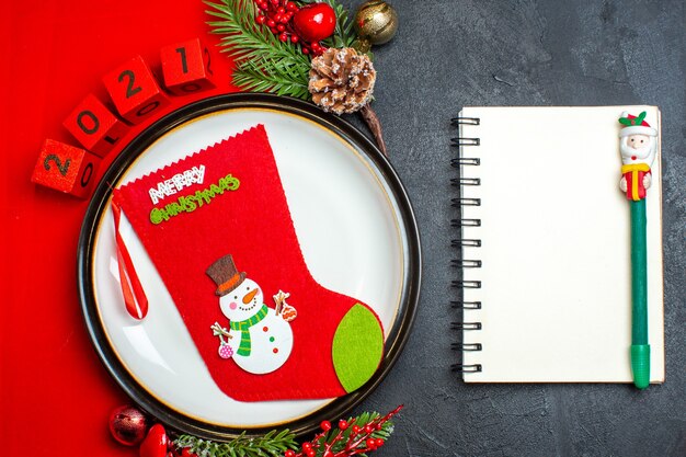 Vista aérea del fondo de año nuevo con calcetín de Navidad en accesorios de decoración de plato de cena ramas de abeto y números en una servilleta roja junto al cuaderno con bolígrafo sobre una mesa negra