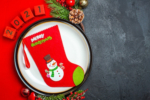 Vista aérea del fondo de año nuevo con calcetín de Navidad en accesorios de decoración de plato de cena ramas de abeto y números en una servilleta roja sobre una mesa negra