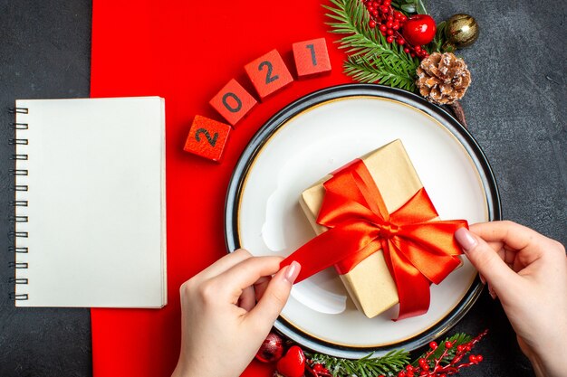 Vista aérea de fondo de año nuevo con regalo en plato de cena accesorios de decoración ramas de abeto y números en una servilleta roja y cuaderno sobre una mesa negra