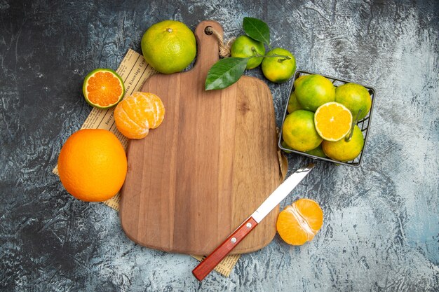 Vista aérea de frutas cítricas frescas con hojas sobre una tabla de cortar de madera cortada en mitades y un cuchillo en un periódico sobre fondo gris