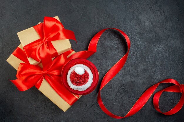 Vista aérea de hermosos regalos con cinta roja y sombrero de santa claus en mesa oscura