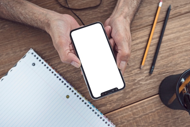 Una vista aérea de la mano del hombre que sostiene el teléfono celular con pantalla blanca sobre el escritorio de madera