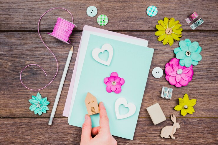Foto gratuito una vista aérea de la mano humana que hace la tarjeta de felicitación con las flores y el bloque de madera de la casa en el escritorio