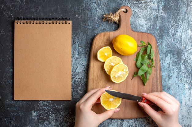 Vista aérea de una mano para picar limones frescos y menta en una tabla de cortar de madera junto al cuaderno espiral sobre fondo oscuro