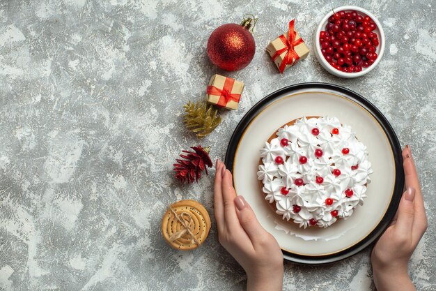 Vista aérea de la mano que sostiene un delicioso pastel con crema de grosellas en un plato y cajas de regalo apiladas galletas conos de coníferas sobre fondo gris