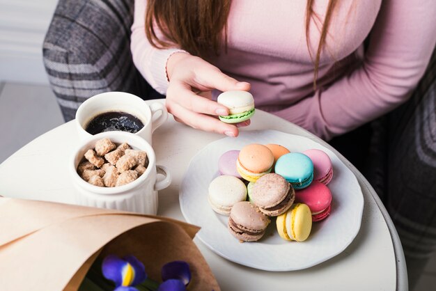 Una vista aérea de la mano que sostiene los macarrones con los cubos del azúcar marrón y la taza de café en la tabla blanca