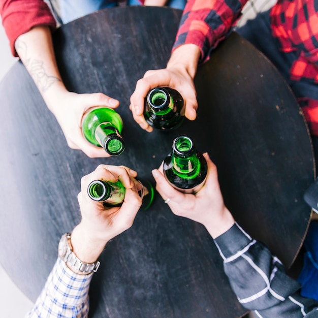 Vista aérea de las manos de un amigo sosteniendo las botellas de cerveza verde en una mesa de madera negra