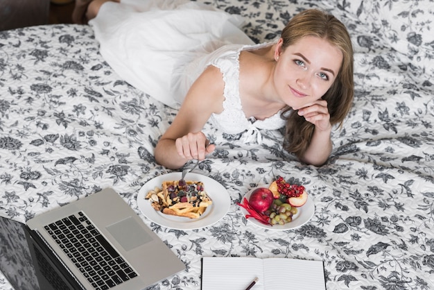 Una vista aérea de una mujer joven acostada en la cama insertando un tenedor en el desayuno de waffle mirando a la cámara