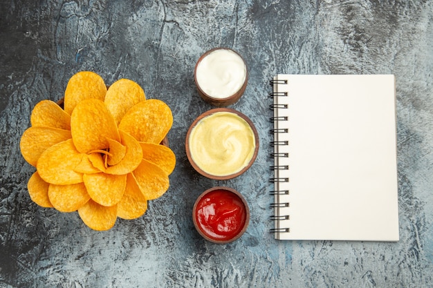 Vista aérea de papas fritas caseras decoradas con forma de flor y sal con mayonesa de ketchup y cuaderno sobre mesa gris
