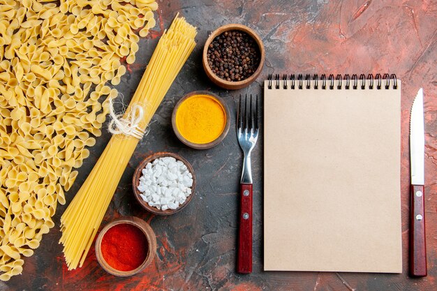 Vista aérea de pasta cruda en diversas formas diferentes especias y cuaderno con tenedor y cuchillo en el cuadro negro