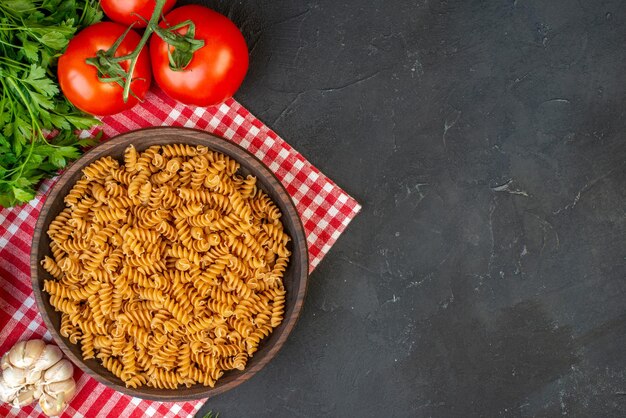 Vista aérea de pastas crudas en una tabla de cortar de madera sobre una toalla roja pelada y tomates ajos verdes en el lado derecho sobre fondo oscuro