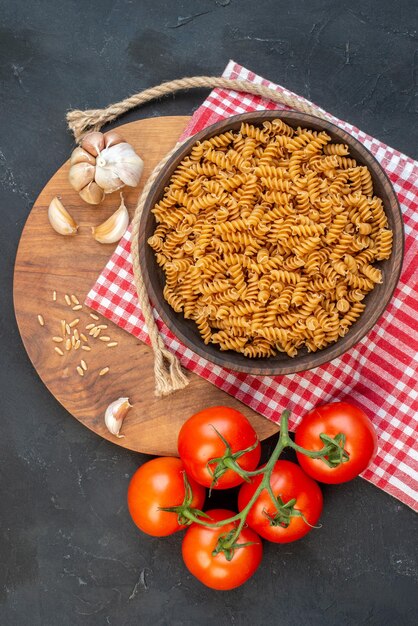 Vista aérea de pastas crudas en un tazón marrón sobre una toalla roja pelada, ajos, arroz sobre una tabla redonda de madera, tomates, cuerda sobre una mesa negra.