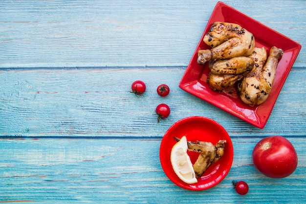 Vista aérea de patas de pollo asadas y alitas de pollo en placa roja con tomates y rodaja de limón contra el escritorio de madera azul