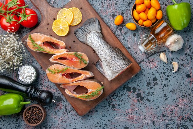 Vista aérea de pescados crudos, rodajas de limón, pimienta verde sobre tabla de cortar de madera y botella de aceite de verduras, kumquats en la tabla de colores azul negro